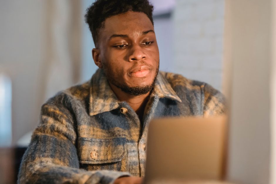 Concentrated young bearded African American man in trendy shirt sitting in front of laptop and attentively looking at screen